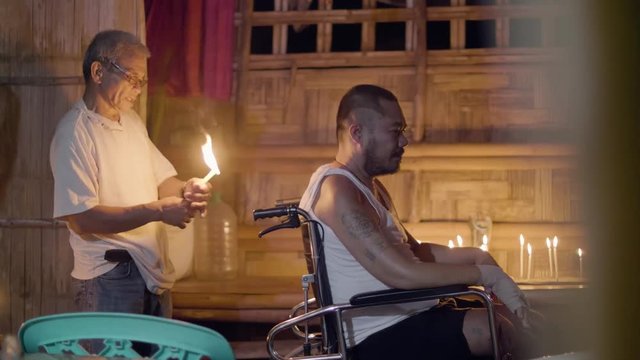 Healer burning candle during magical ritual to treatment sick man in wheelchair in magic house. Traditional asian healing ritual. Ethnic authentic traditions.