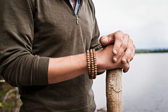 Mid section of man with bracelet standing by lake and leaning against wooden pole 