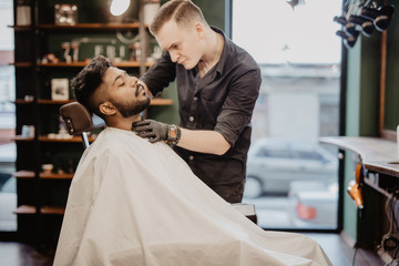 Grooming of real man. Young indian bearded man getting beard haircut at hairdresser while sitting in chair at barbershop