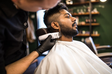 Perfect trim at barbershop. Young bearded man getting haircut by hairdresser with electric razor while sitting in chair at barbershop