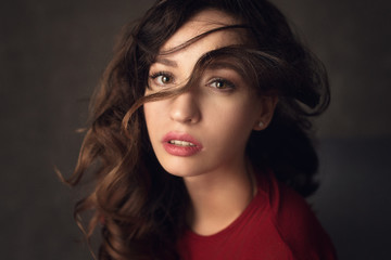 Fototapeta premium beautiful woman with brunette hair in red dress with curl on her face and red lips posing on the dark background in studio