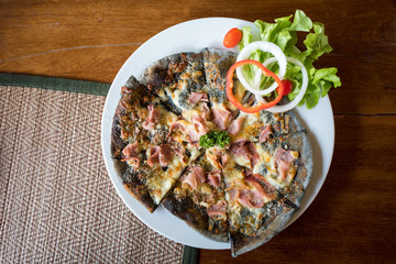 Homemade Pizza ham cheese with charcoal dough decorated with fresh vegetables on white round plate.