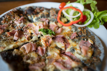 Homemade Pizza ham cheese with charcoal dough decorated with fresh vegetables on white round plate.