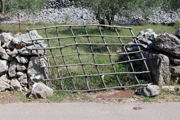 Tilted improvised backyard wooden doors made from wooden sticks supported with makeshift stone wall with tall grass and olive trees in background on warm sunny day