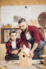Young caucasian father and his pre-teen son working together in a wooden workshop, building a wooden bird house. Little boy dressed in apron and wearing protective glasses working with a hummer