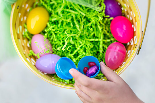 Close Up View Of An Open Plastic Egg With Easter Candy In It. A Hand Is Pulling Out Candy Out Of The Easter Basket With Other Eggs And Paper Grass In It