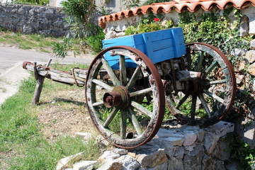 Vintage retro cracked dilapidated part of wooden horse carriage with metal wheel frames used as garden decoration for planting flowers in front of traditional stone wall with red roof tiles on warm su