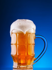 Faceted mug of the light beer with foam on blue background
