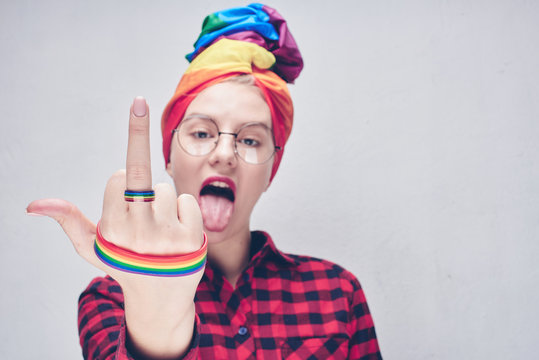 A beautiful young woman with glasses in a rainbow turban and a red shirt shows her middle finger and tongue against a white wall. LGBTQ Lesbians, gays, bisexuals, transgender, queer. Fuck you concept