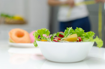 healthy fresh mix vegetable salad in white bowl with delicious glazed donuts in background on desk in kitchen interior at house, vegetarian, lifestyle, vegan, good healthy, diet food and drink concept