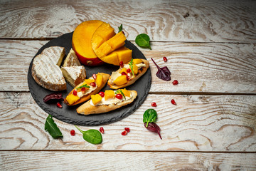 Impressive Cheese Plate or cheese platter snacks for Wine. Brushetta with mango, Camembert cheese and pomegranate served on a shale board, rustic wooden table. Top view