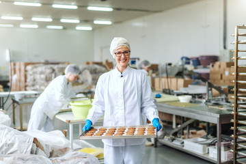 Smiling blonde Caucasian employee in sterile uniform and with eyeglasses standing and holding tray with cookies. Food factory interior. In background other employees working.