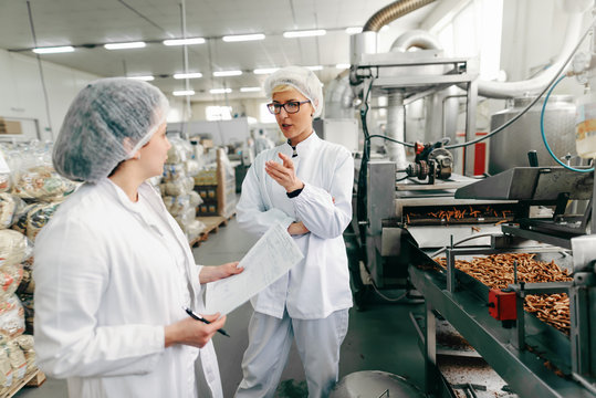 Coworkers in white uniforms and with sterile caps on heads discussing about quality of products while standing in food factory.