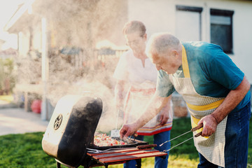 Grandparents with aprons grilling meat and vegetables on sticks while standing in backyard at spring.