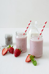Strawberry smoothie, milk shake. 2 jars of strawberry milk smoothie and 1 jar of chia seeds on white table board, embellished with real strawberries and fresh mint. Glass pitcher with milk in the back