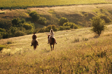 Male instructor and girl galloping on brown horses in the field at sunset