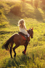 Girl blonde rides a brown horse in a field at sunset