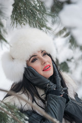 Woman with white fur cap and waistcoat enjoying the winter scenery near an iron fence. Attractive long hair brunette girl posing in winter landscape. Beautiful female with red lips in bright cold day