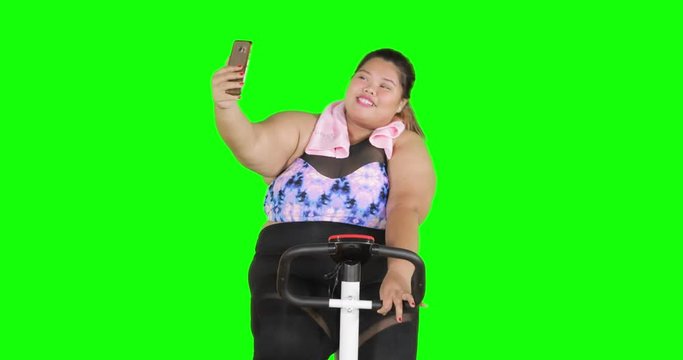 Overweight woman exercising on exercise bike while taking selfie photo. Shot in 4k resolution with green screen background