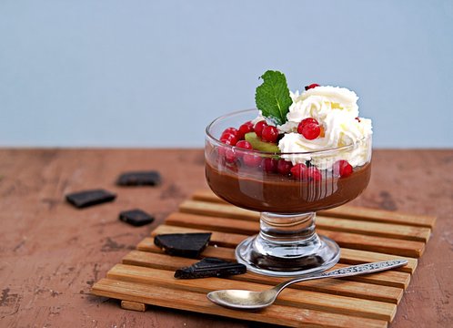 Chocolate pudding with whipped cream, red currants and kiwi in a glass ice-cream bowl on a brown concrete background.