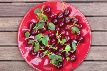 Red plate of ripe cherries with green leaves on a wooden table