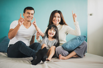 Happy Cheerful smiling Young Family mother father or parent and daughter sitting on the white bed at home and the hand pointing up, blue background