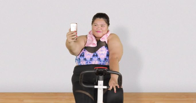 Overweight woman taking selfie photo while exercising on exercise bike at home. Shot in 4k resolution