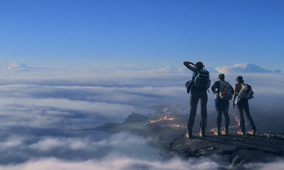 Three hikers on top above clouds at evening soft light watching valley with city lights