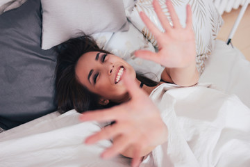 Obraz na płótnie Canvas Happy smiling beautiful long hair asian girl young woman lying down in bed and pulling hands towards camera, cozy morning