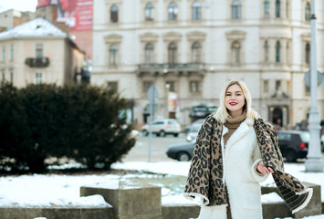 Cheerful blonde model wears winter coat and knit scarf walking at the city. Empty space