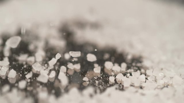 SLOW MOTION, MACRO, DOF: White sugar is being sprinkled over the granite kitchen counter. Pristine rock salt is scattered around the stone kitchen table. Sea salt is sprinkled over the black surface.