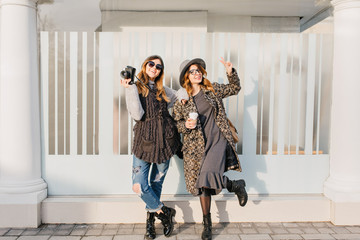Two fashionable joyful smiling girls having fun on sunny street in city. Stylish look, travelling together, wearing modern trend clothes, walking with coffe to go, expressing positive emotions © Look!