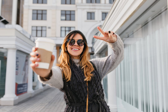 Expressing brightful positive emotions of fashionable city girl stretching coffee to go to camera on sunny street. Beautiful smiling girl in modern sunglasses, knitted hat having fun outdoor