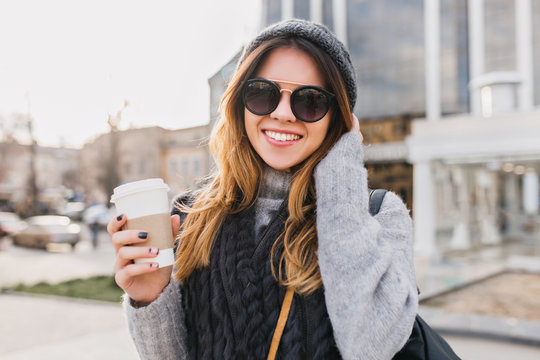 Portrait fashionable urban young woman with coffee to go walking in sunny city centre. Amazing smiling girl in modern sunglasses, knitted hat, woolen sweater having fun outdoor