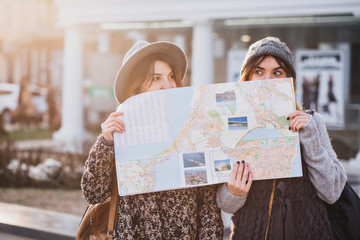 Funny positive image of fashionable girls on sunny street having fun in city, hiding behind the citymap. Travelling together, best friends, get a lost in big city, true emotions