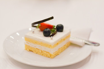 Piece of square vanilla cake with blueberry, strawberry and chocolate on white dish and table in wedding party.
