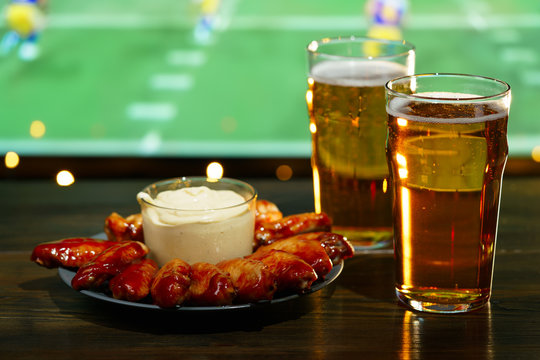 Hot barbecue chicken wings with 2 beer glasses on a dark wooden table served with honey mustard sauce. Football on a background, high resolution