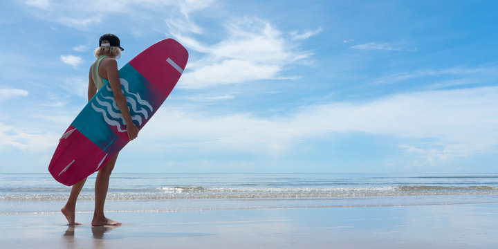 Woman with pink blue board standing  on a beach blue sky clouds copy space