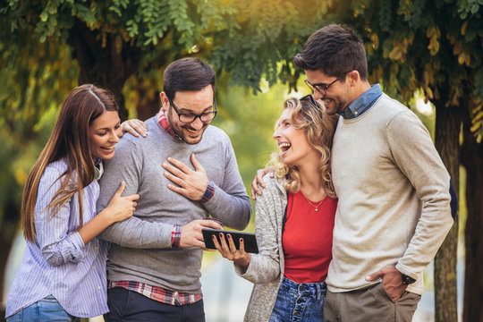 Four happy smiling young friends walking outdoors in the park holding digital tablet