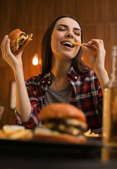 Young woman eating French fries and tasty burger in cafe