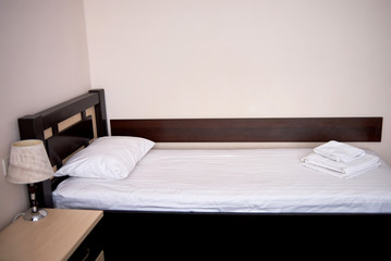 Hotel bedroom interior with empty single bed with wooden headboard and bedside table near beige wall, copy space. White sheet, soft pillow and towels on bed