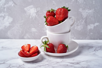 Red organic strawberries in white ceramic cup and strawberry slices in bowl on gray concrete background, copy space. Healthy food concept, still life