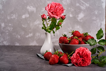 Red ripe strawberries in ceramic bowl on linen table napkin and pink rose in vase on gray concrete background, copy space. Healthy food concept, still life