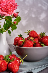 Red ripe strawberries in ceramic bowl on linen table napkin and pink rose in vase on gray concrete background, copy space. Healthy food concept, still life