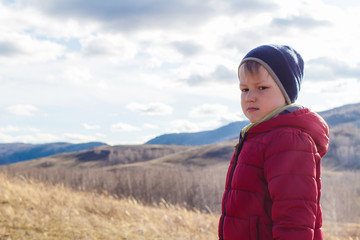 serious six-year-old boy in a warm red jacket and hat on a background of clouds outdoors in spring