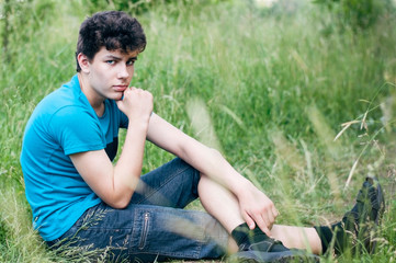 Naklejka na ściany i meble A beautiful but sad teenager sits on the grass with a thoughtful look. A boy thinks how to make peace with his parents, with whom he recently quarreled