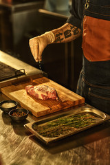 Hand of professional chef in apron and white glove salting raw meat. Beef steak on wooden cutting...
