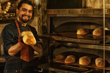 Smiling baker showing fresh baked bread and holding it in his hand. Beautiful and handsome man standing near special convection oven in bakery. Concept of baker's shop. - obrazy, fototapety, plakaty