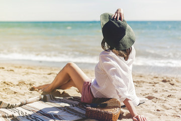 Summer vacation concept. Happy young woman relaxing on beach. Hipster slim girl in white shirt and hat sitting and tanning on beach near sea with waves, sunny warm weather. Peaceful calm moment - Powered by Adobe