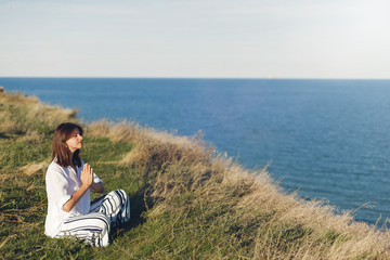 Naklejka na ściany i meble International Yoga day concept. Young beautiful woman practicing yoga on beach, sitting in grass. Hipster girl doing yoga, relaxing and listening to sea waves. Meditation, calm, happiness.
